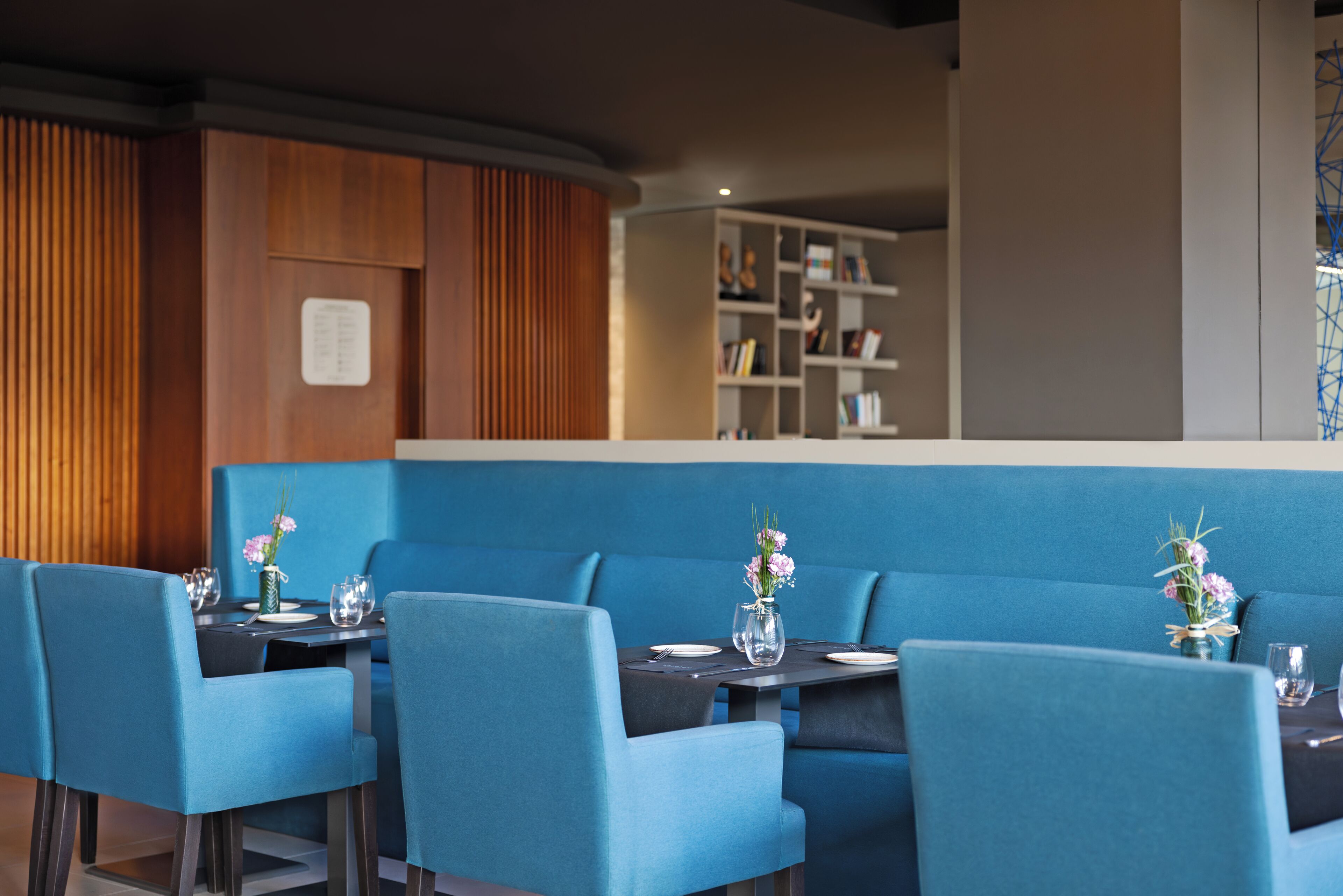 A restaurant with blue upholstered booths, a wooden accent wall, and a bookshelf in the background.