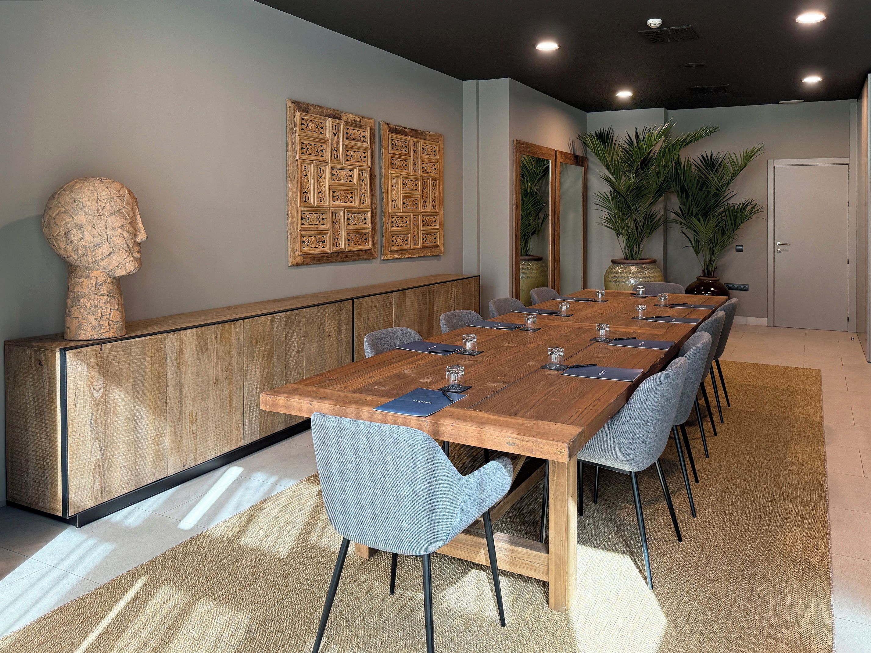 A conference room with a long wooden table, grey upholstered chairs, and a decorative bust on a wooden cabinet.