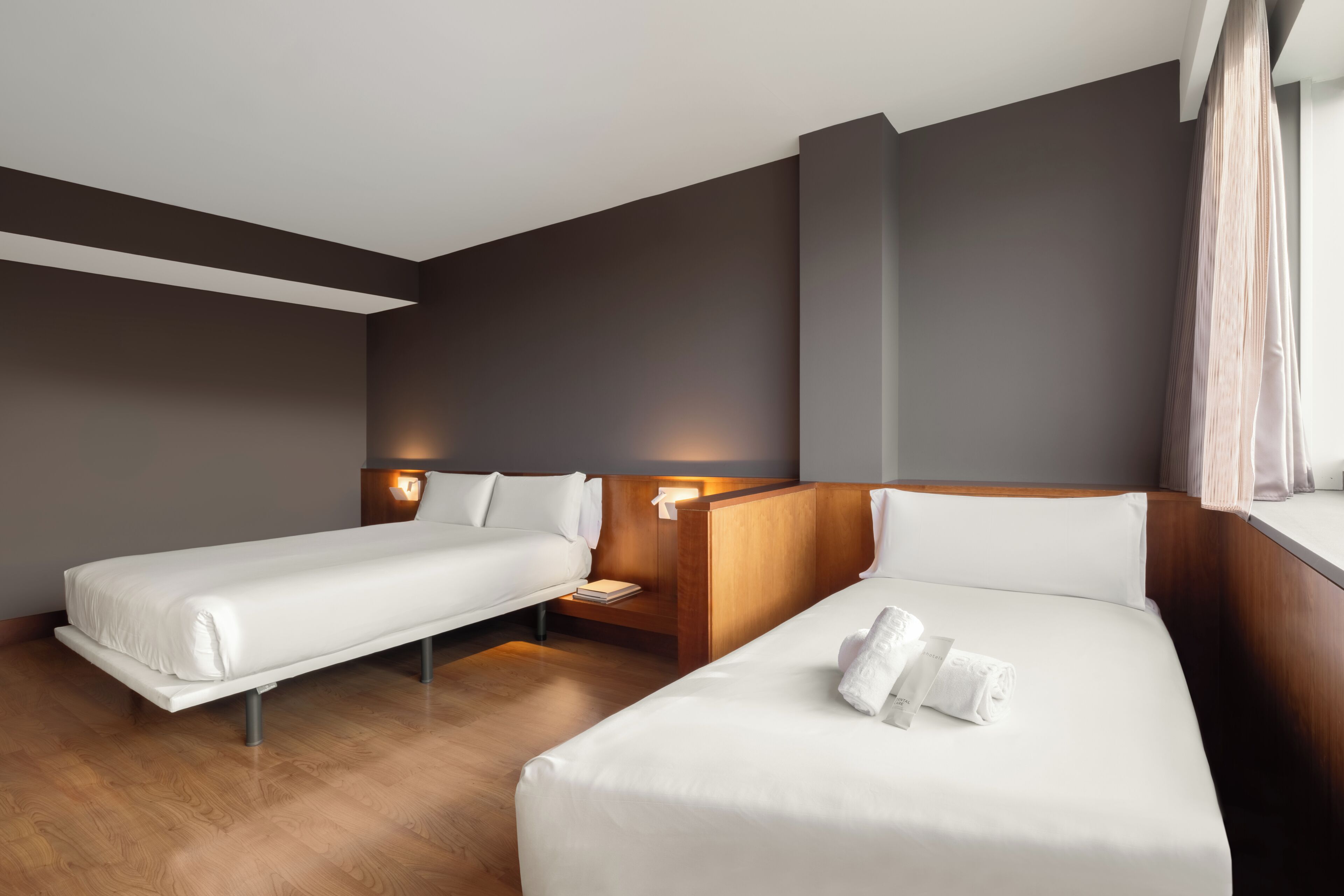 A hotel room with two beds, wooden headboards, a nightstand, and a window with curtains.