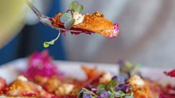 A close-up of a dish with a fork lifting a piece of food, garnished with greens and a pink element, on a plate with other food items and a bread roll in the background.