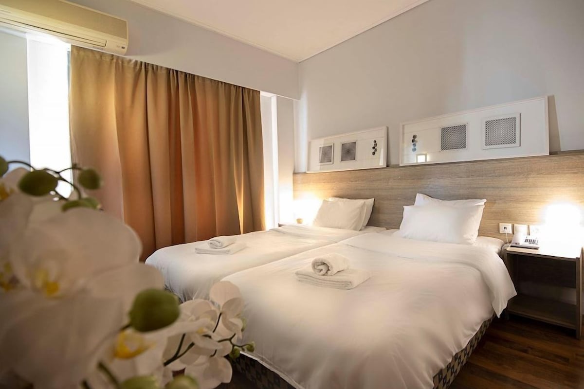 Quarto de hotel com duas camas, cabeceira de madeira, mesa de cabeceira e janela com cortinas.