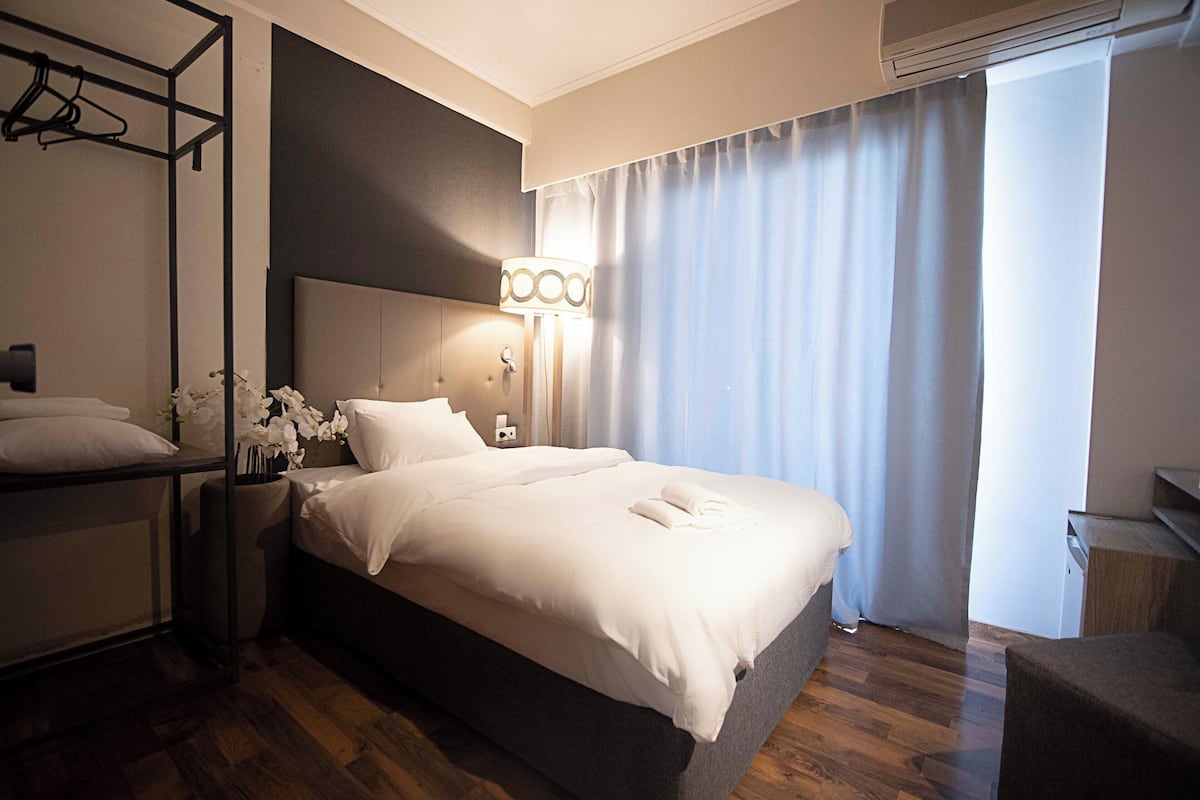 Habitación de hotel con cama, mesita de noche, lámpara, espejo, armario y ventana con cortinas.