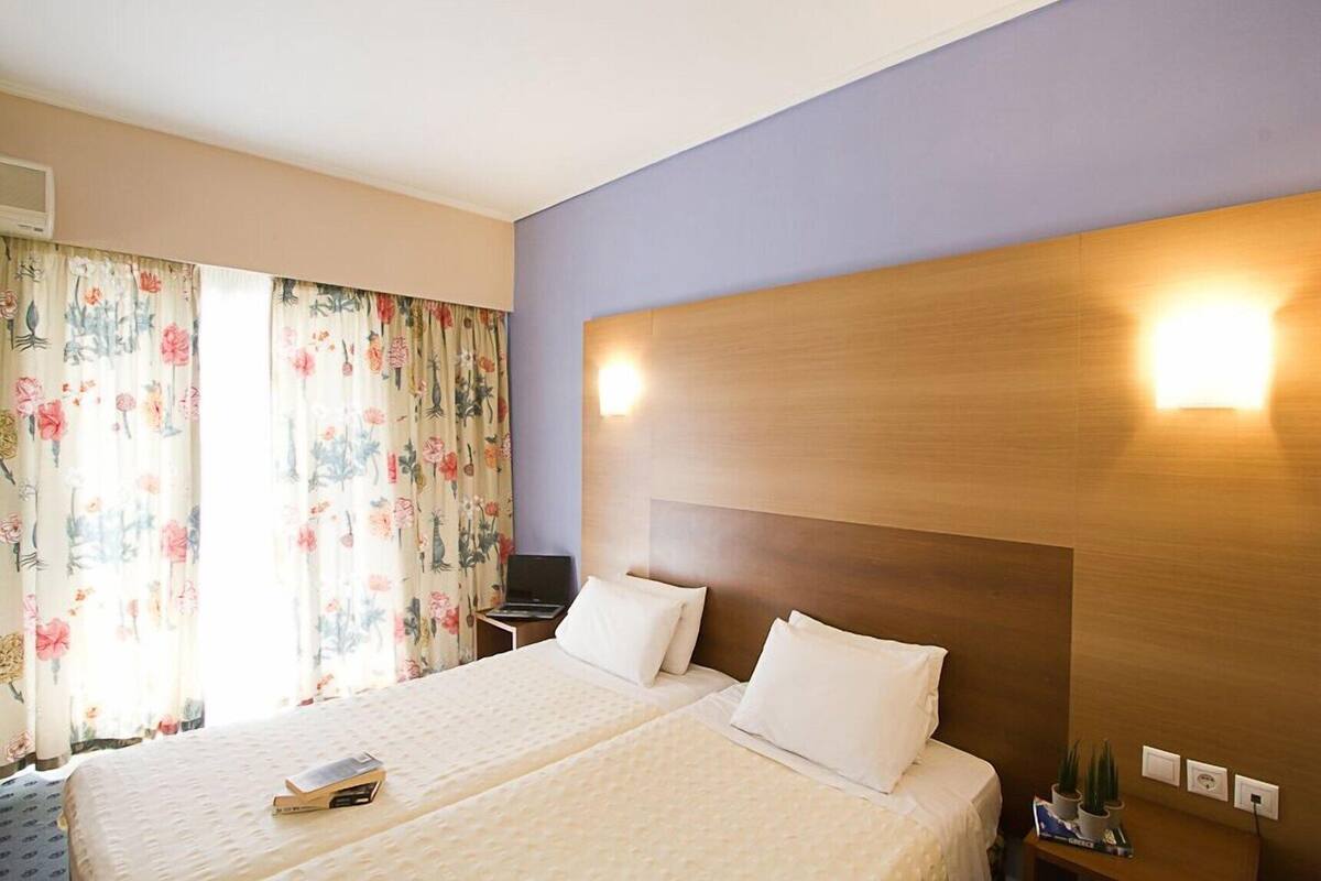 A hotel room with a double bed, floral curtains, a wooden headboard, bedside lamps, a nightstand, and a small table with a plant.