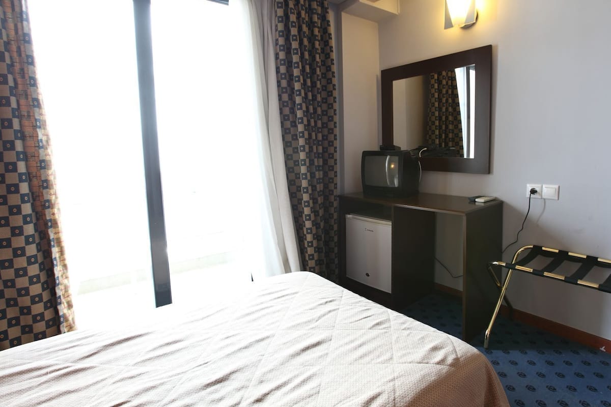 Una habitación de hotel con cama, escritorio con computadora, espejo y ventana con cortinas.