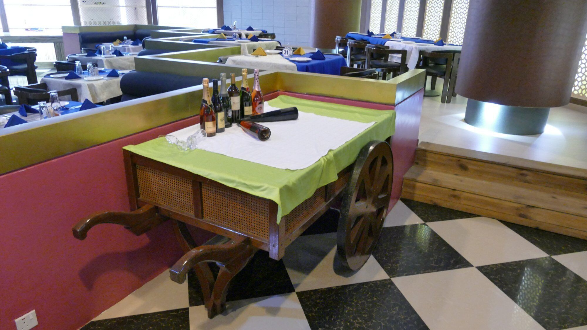 Un restaurante con cabinas y mesas, algunas con servilletas azules. Hay botellas sobre una mesa, y una gran estructura metálica al fondo.