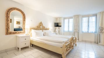 Un dormitorio con una cama grande y ornamentada, una mesita de noche blanca con cajones, un espejo grande y un televisor en la pared.