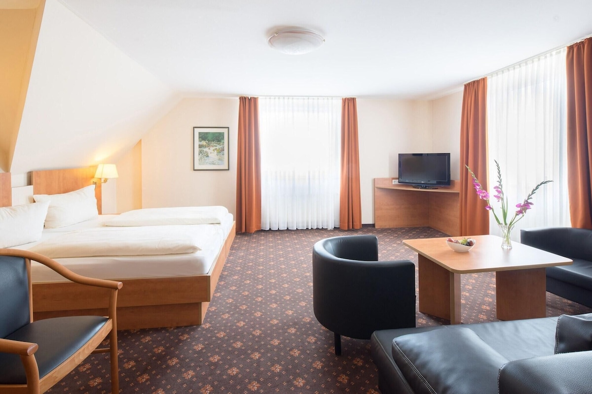 Une chambre d’hôtel avec un lit, deux chaises, une petite table, un téléviseur et une fenêtre avec des rideaux.