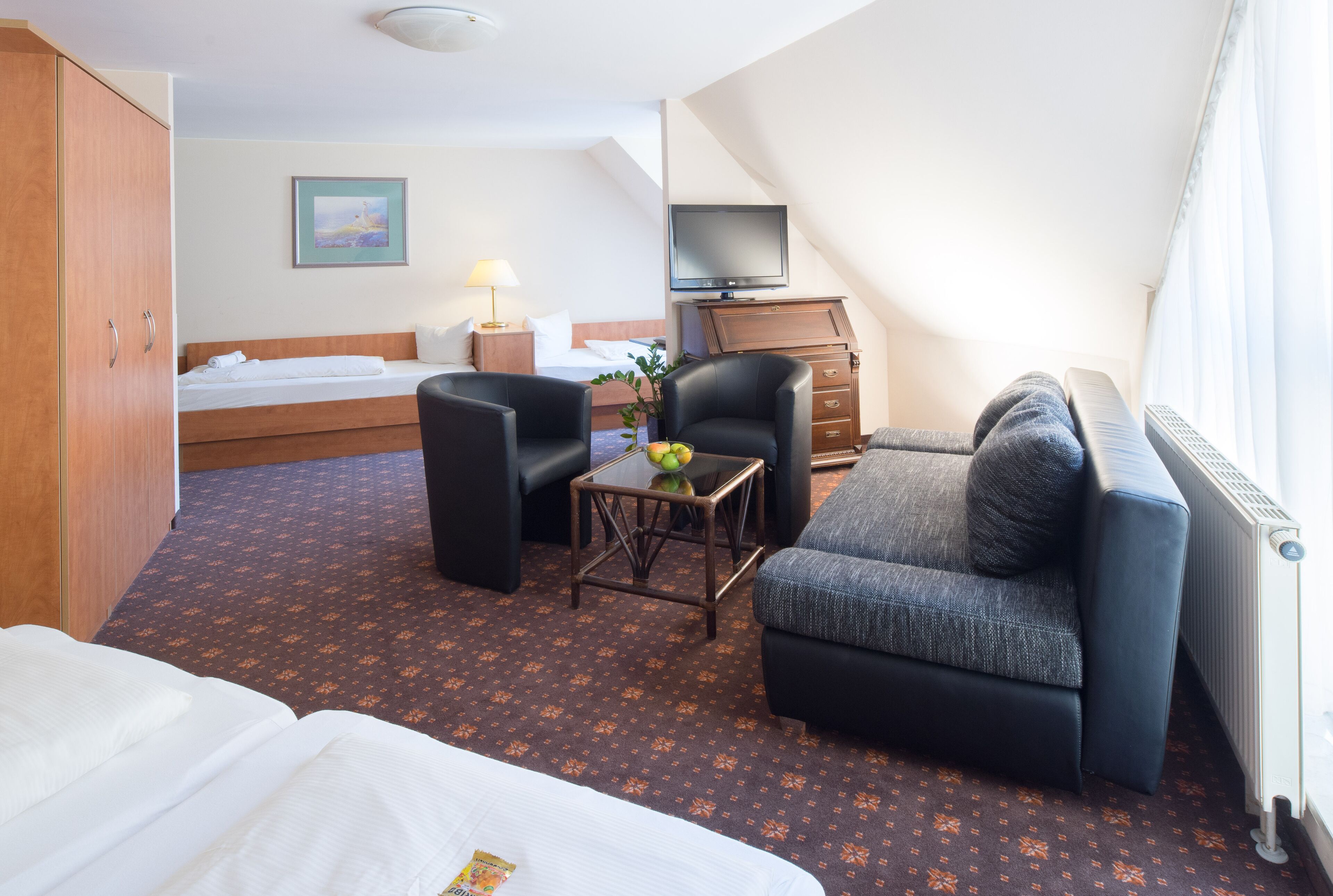 A hotel room with a bed, a sofa, a chair, a small table with a fruit bowl, a television, and a radiator.