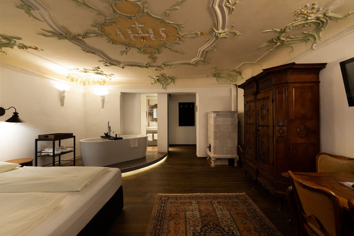 A hotel room with a large bed, a sitting area with a wooden table, a white bathtub, and a wooden wardrobe.