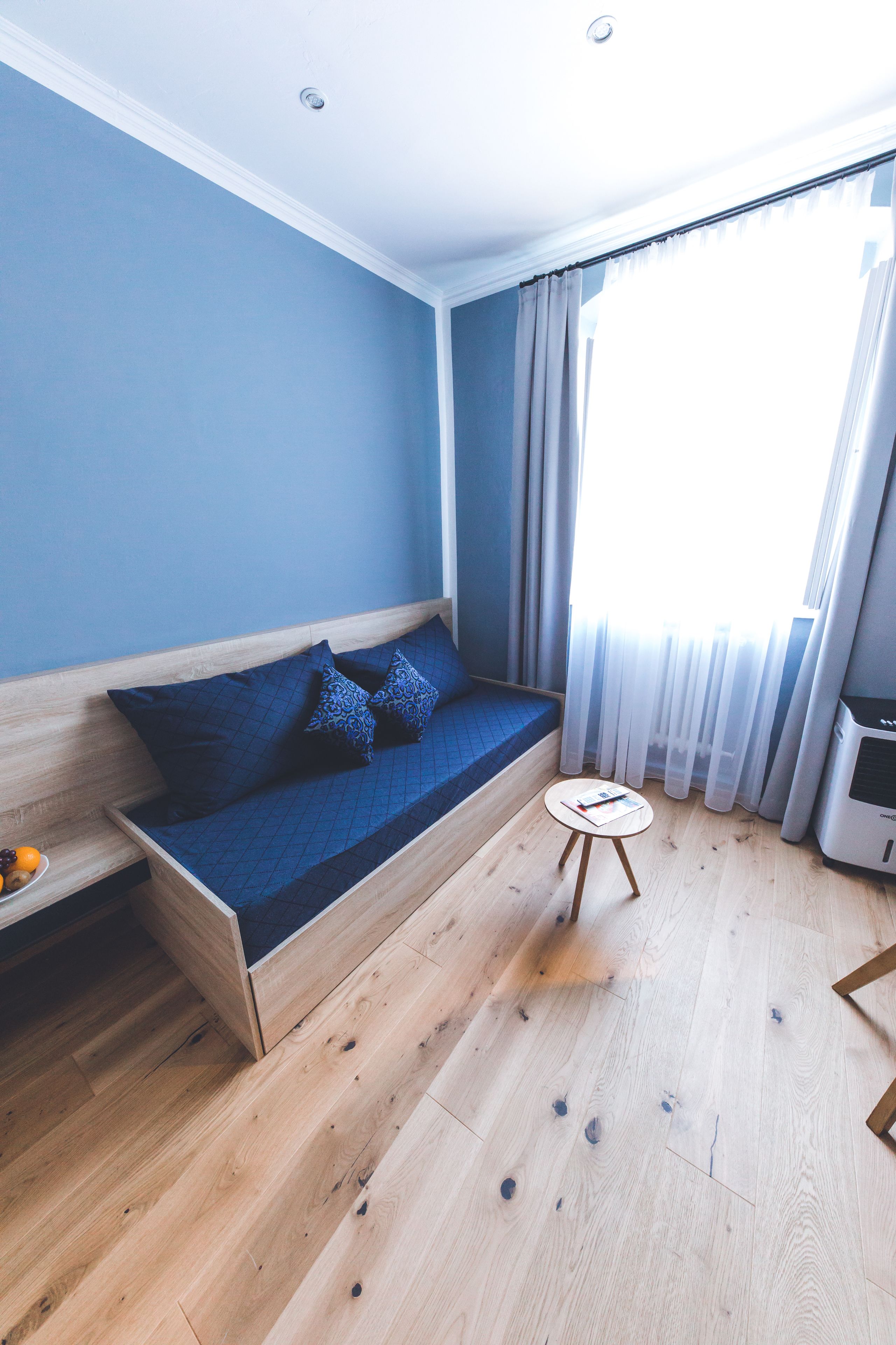 Una habitación con suelo de madera, pared azul, sofá con cojines estampados, una mesita redonda y una ventana con cortinas.