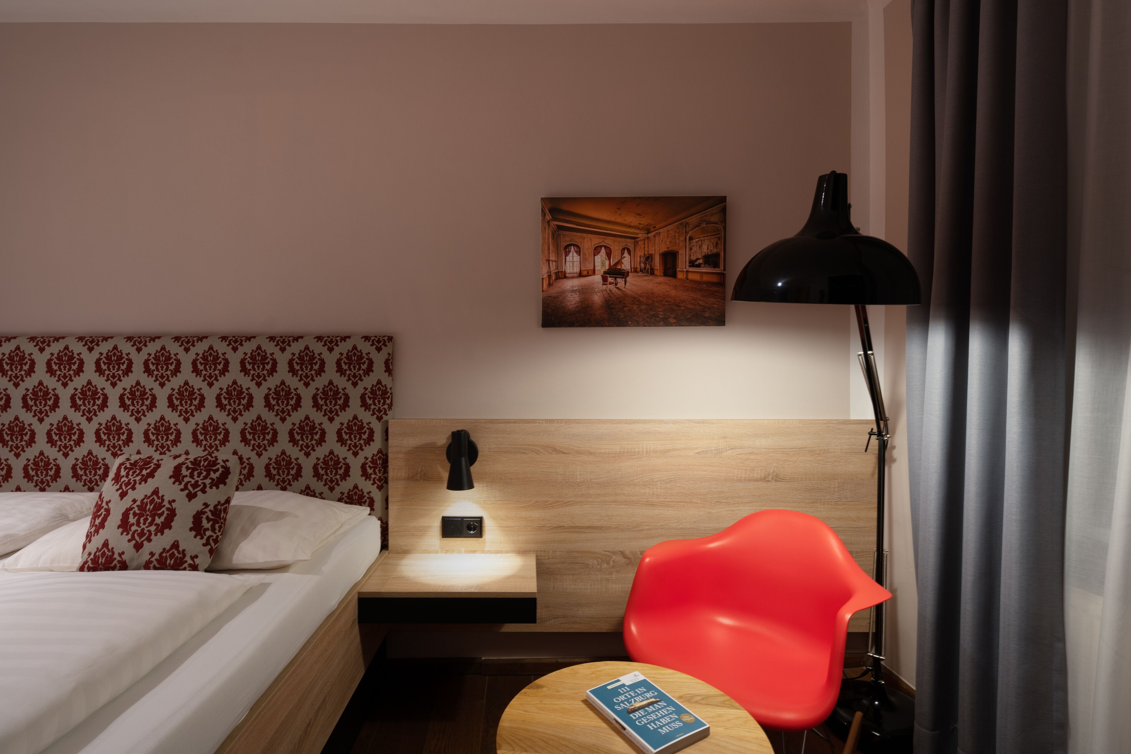 A hotel room with a bed, a nightstand with a lamp, a red chair, a small table with a book, and a painting on the wall.