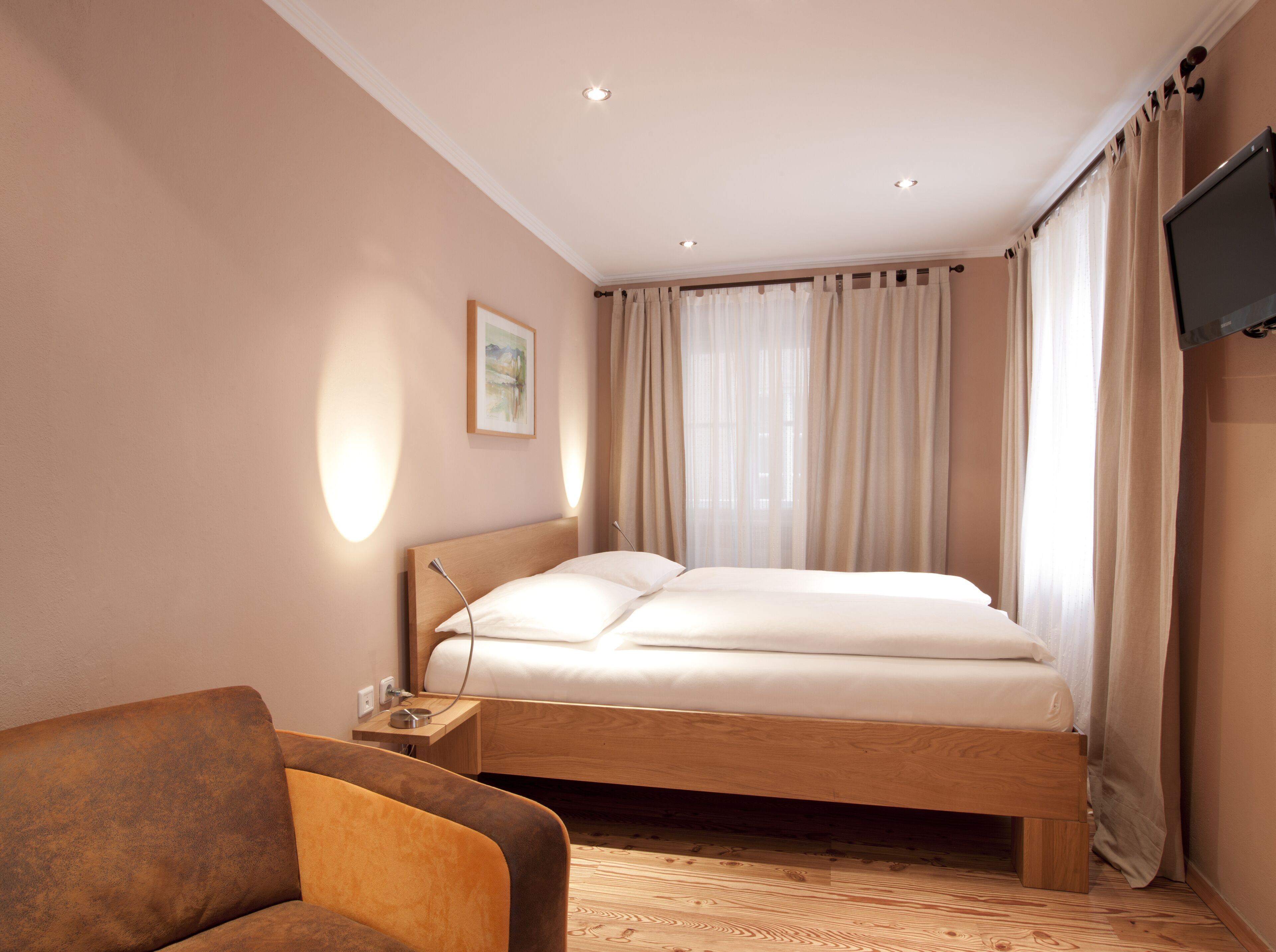 A modern hotel room with a wooden bed, a bedside table, a chair, and a flat-screen TV.