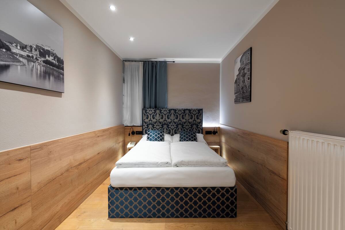 A modern hotel room with a wooden headboard, a large bed, a nightstand, a wall-mounted radiator, and a framed picture on the wall.