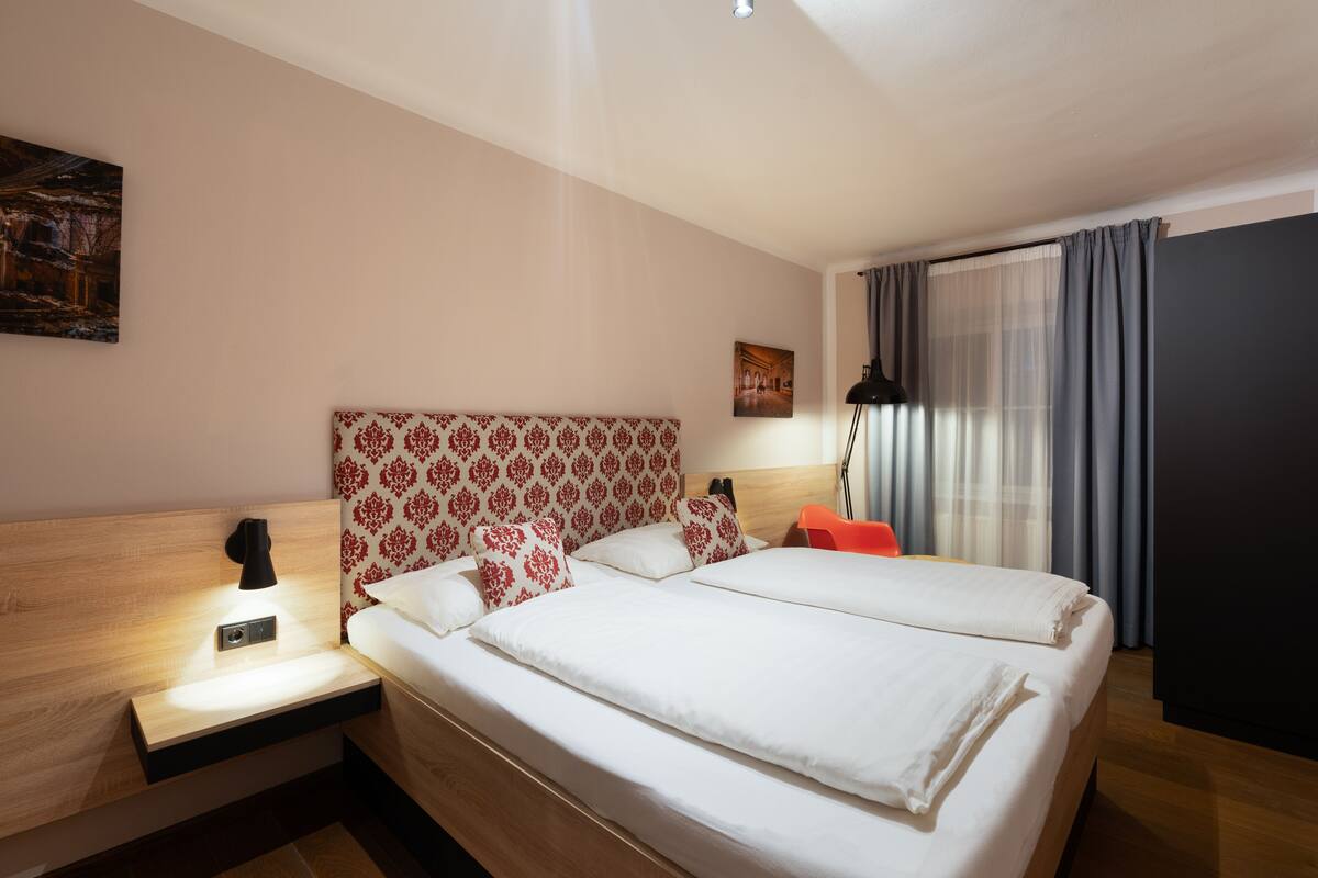 A hotel room with a large bed, a nightstand with a lamp, a headboard with a floral pattern, a painting on the wall, and a window with curtains.