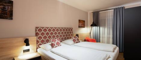 A hotel room with a large bed, a nightstand with a lamp, a headboard with a floral pattern, a painting on the wall, and a window with curtains.