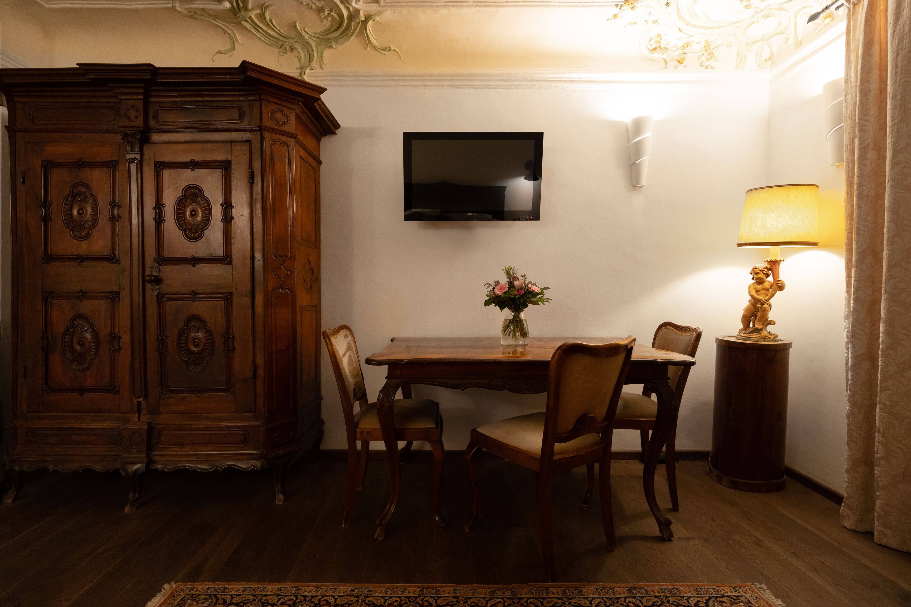 A dining area with a wooden table, chairs, a vase with flowers, a television mounted on the wall, and a lamp on a side table.