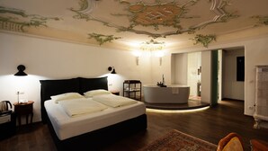 A bedroom with a large bed, a nightstand, a desk, a chair, a bathroom with a bathtub, and a chandelier. - Altstadthotel Kasererbräu (Salzburg)