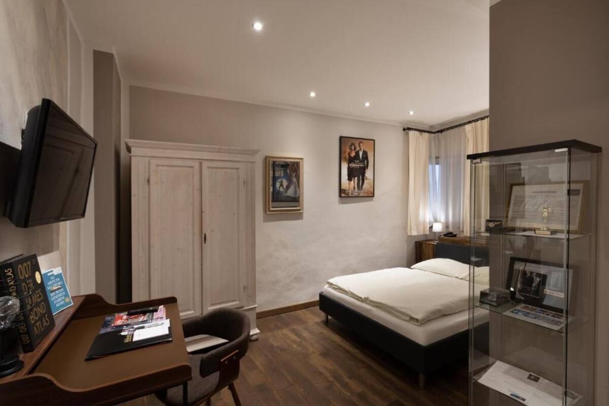A hotel room with a bed, a desk with a chair, a television, a wardrobe, and a glass display case.