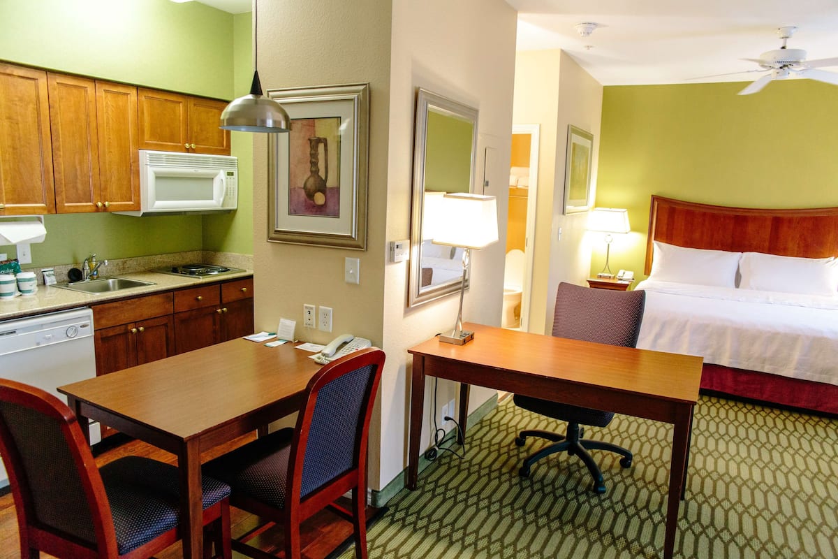 A hotel room with a kitchenette, a dining area with a table and chairs, a bed with a red headboard, and a ceiling fan.