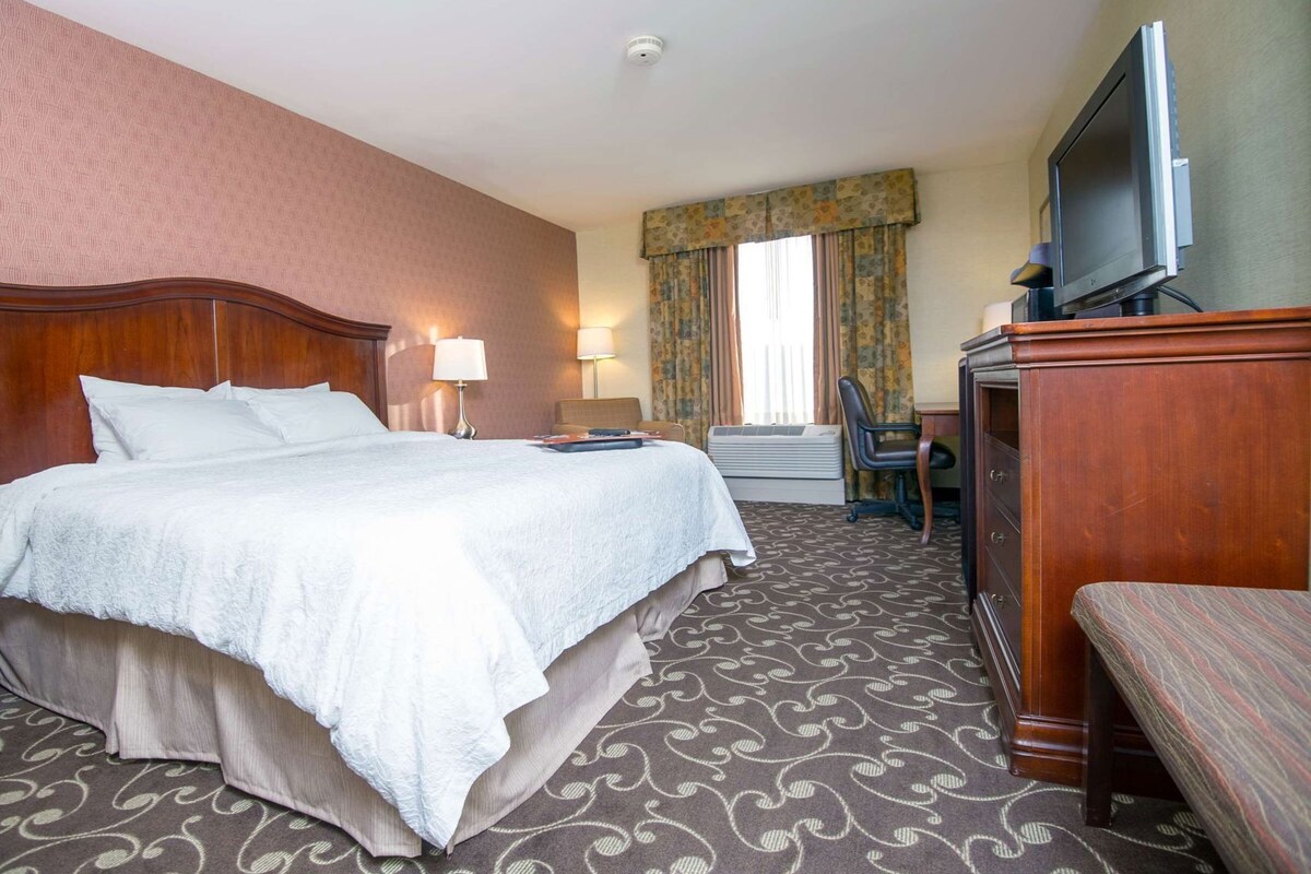 A hotel room with a large bed, a desk with a television, a chair, and a window with curtains.
