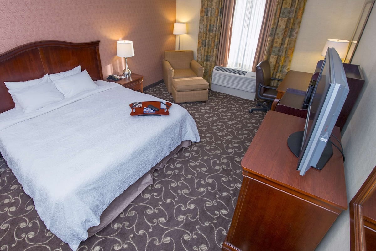 A hotel room with a large bed, a desk with a computer, a chair, a lamp, and an air conditioning unit.