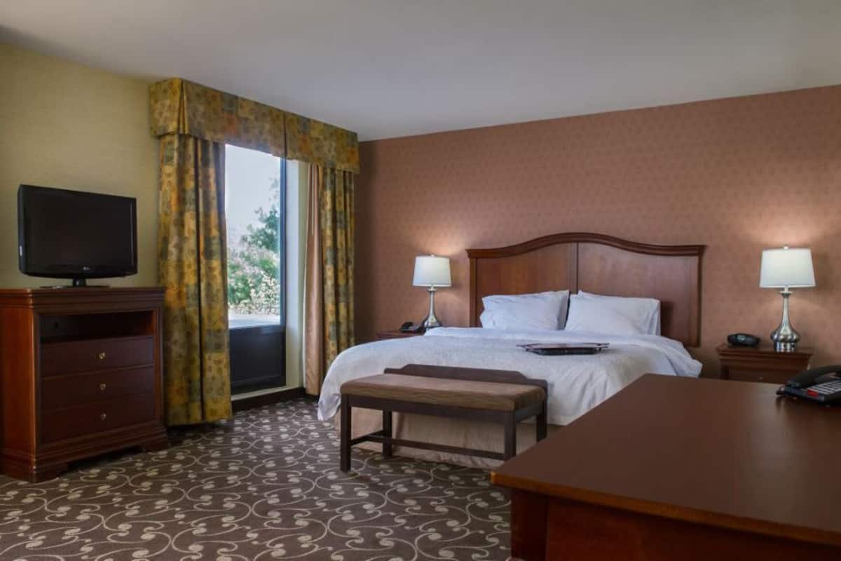 A hotel room with a bed, bedside lamps, a television, a desk, and a window with curtains.