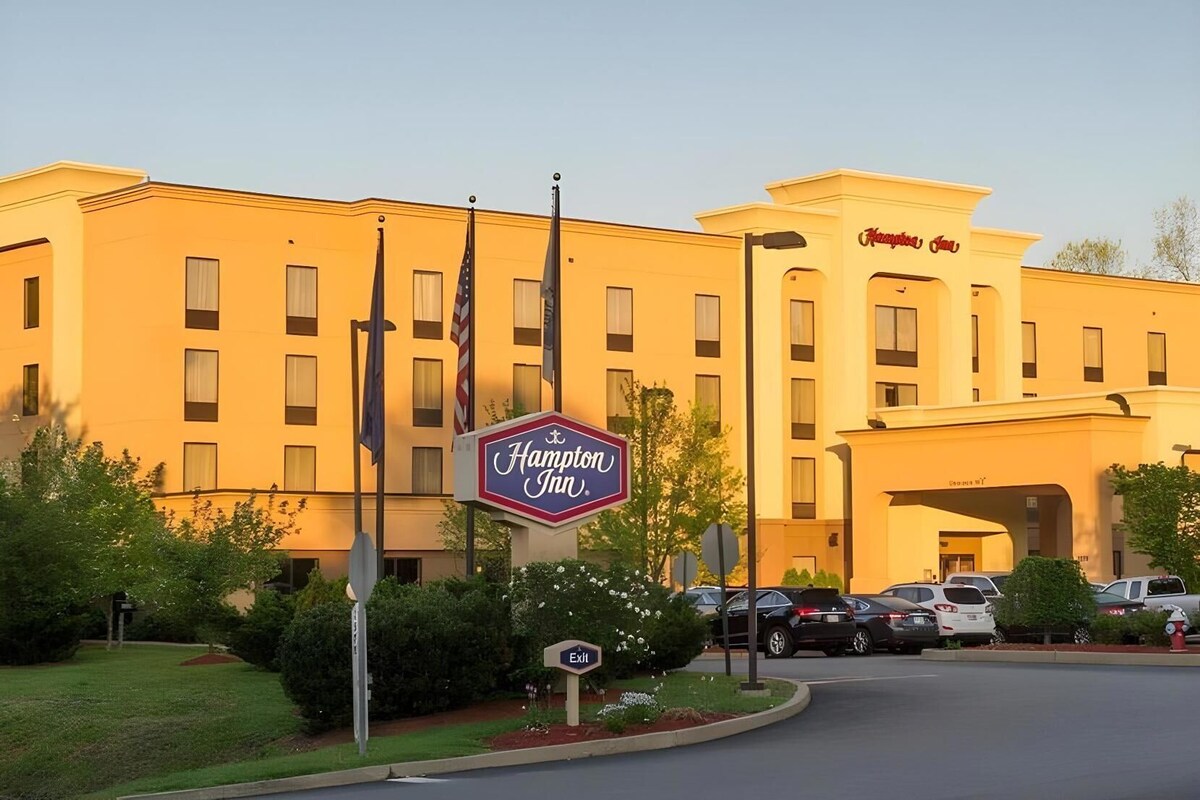 A Hampton Inn hotel with a prominent sign, multiple flags, and a parking lot.