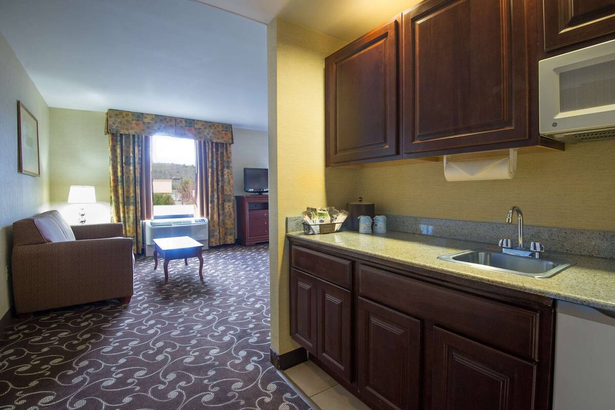 A compact hotel room with a kitchenette, a sofa, a small table, and a window with curtains.