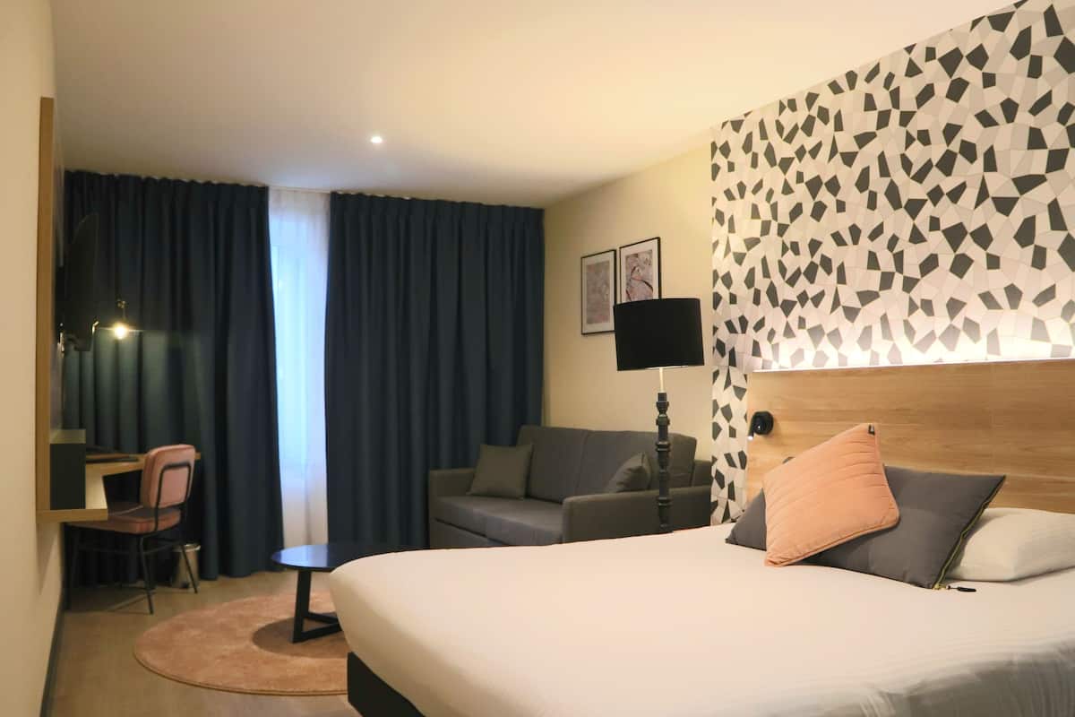 Une chambre d’hôtel comprenant un lit, un bureau, une chaise, un canapé, une lampe et un mur orné d’un motif géométrique.