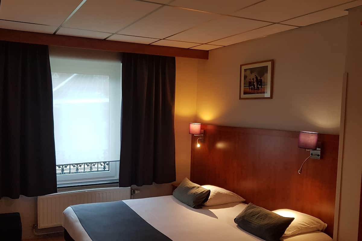 Une chambre d’hôtel avec un lit, une fenêtre avec des rideaux, un téléviseur fixé au mur, une lampe de chevet et un tableau encadré accroché au mur.