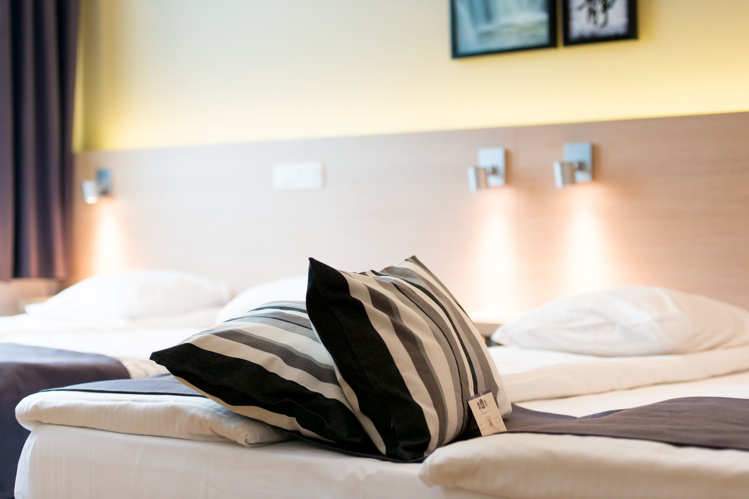 Quarto de hotel com cama bem arrumada, almofadas listradas e cabeceira com luzes fixadas na parede.