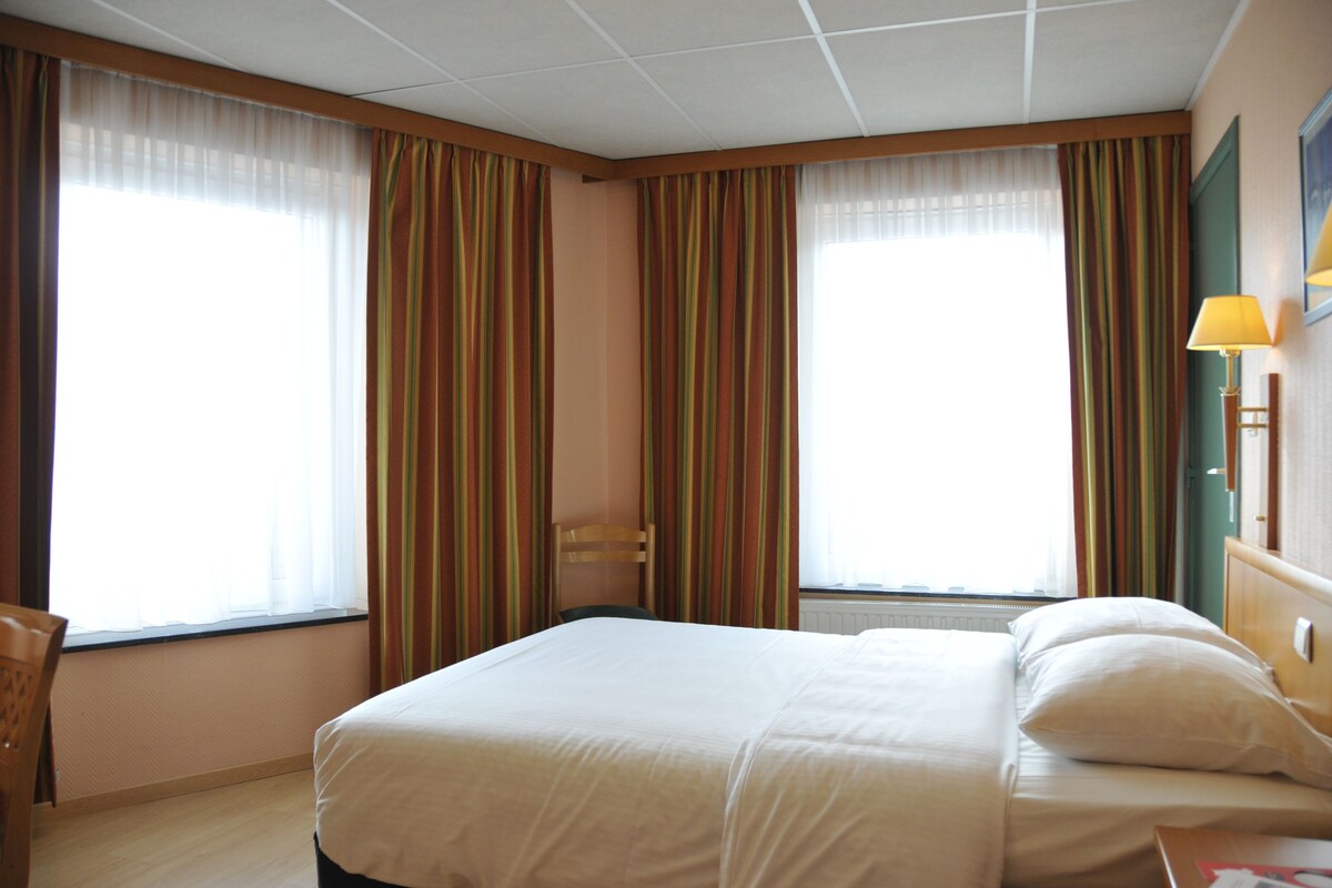 A hotel room with a large bed, two windows with curtains, a chair, and a bedside table with a lamp.