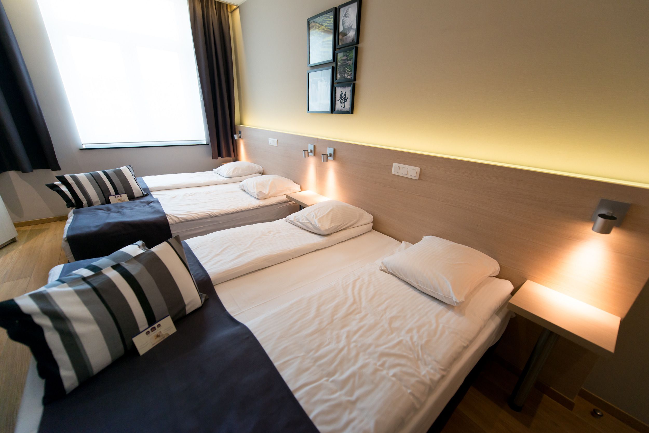 A hotel room with three single beds, each with a pillow and a bedside table with a lamp.