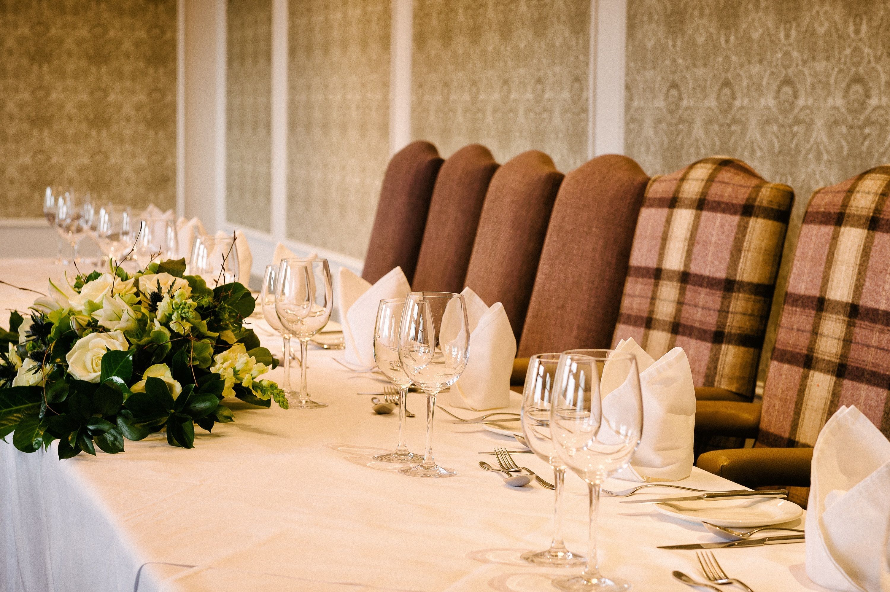 A long dining table with white tablecloth, set with wine glasses, plates, and cutlery, flanked by upholstered chairs with plaid upholstery.