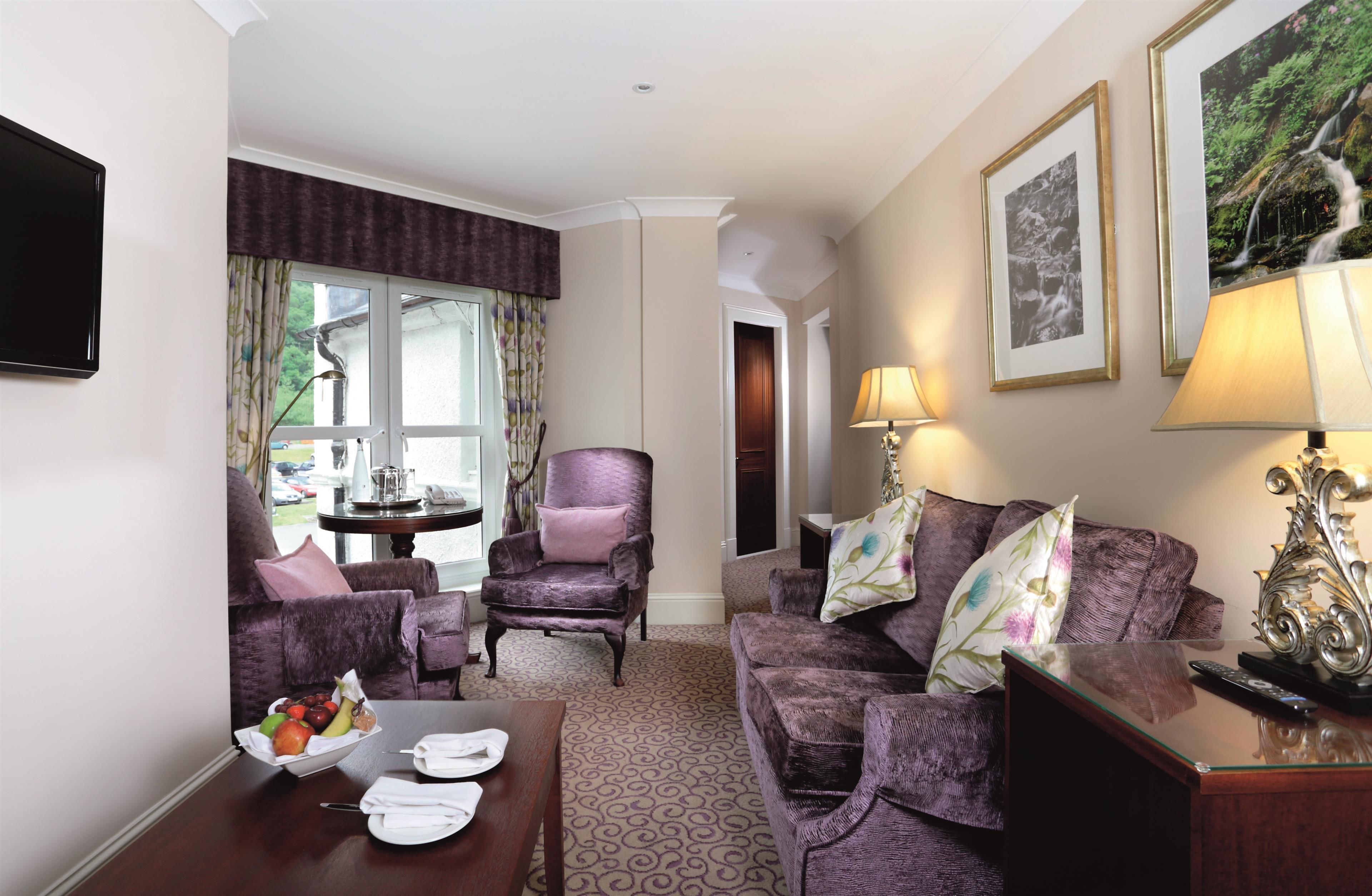 A living room with a purple sofa, a matching armchair, a coffee table with fruit and napkins, and a TV.