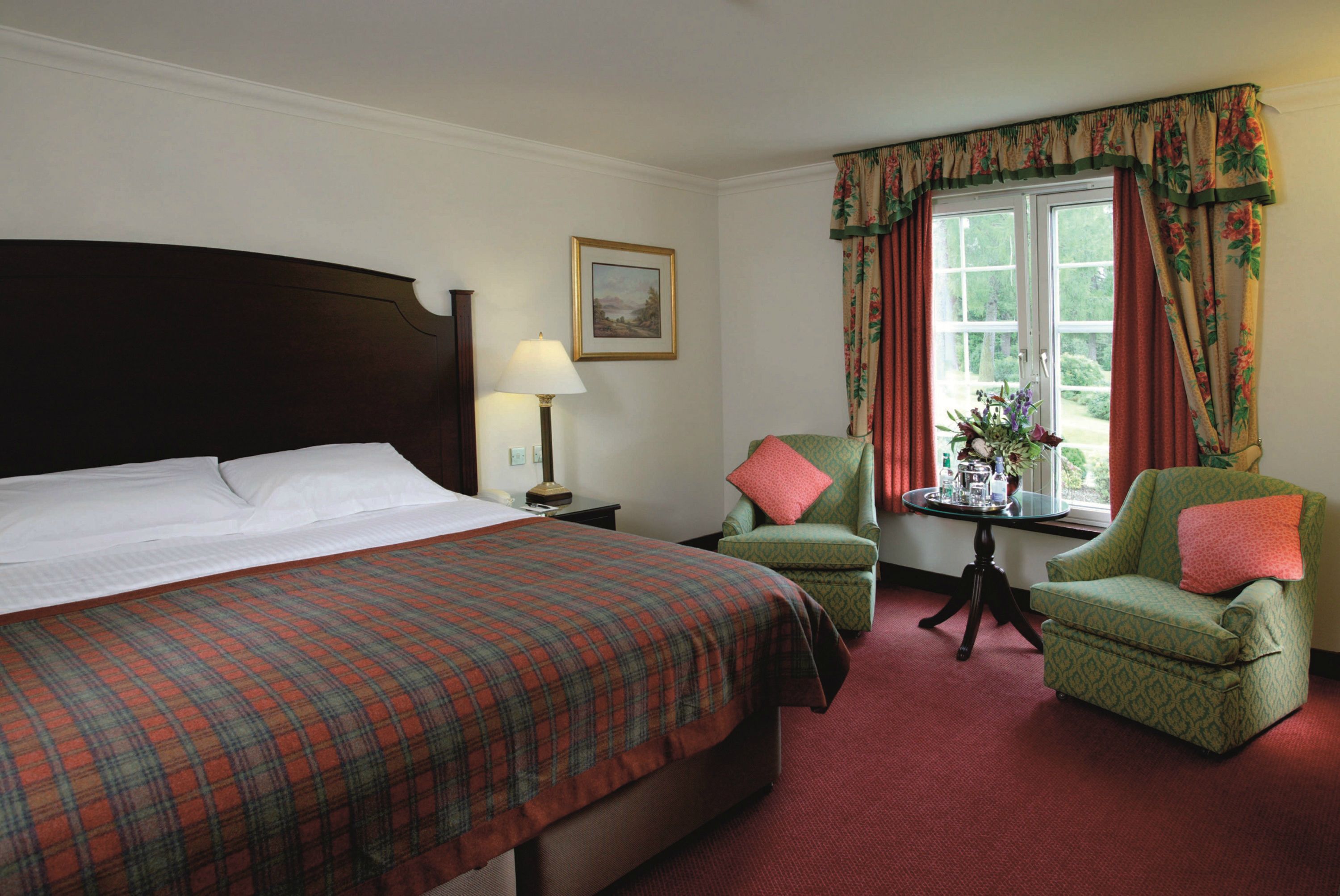 A hotel room with a large bed, two armchairs, a small table with a lamp, and a window with curtains.