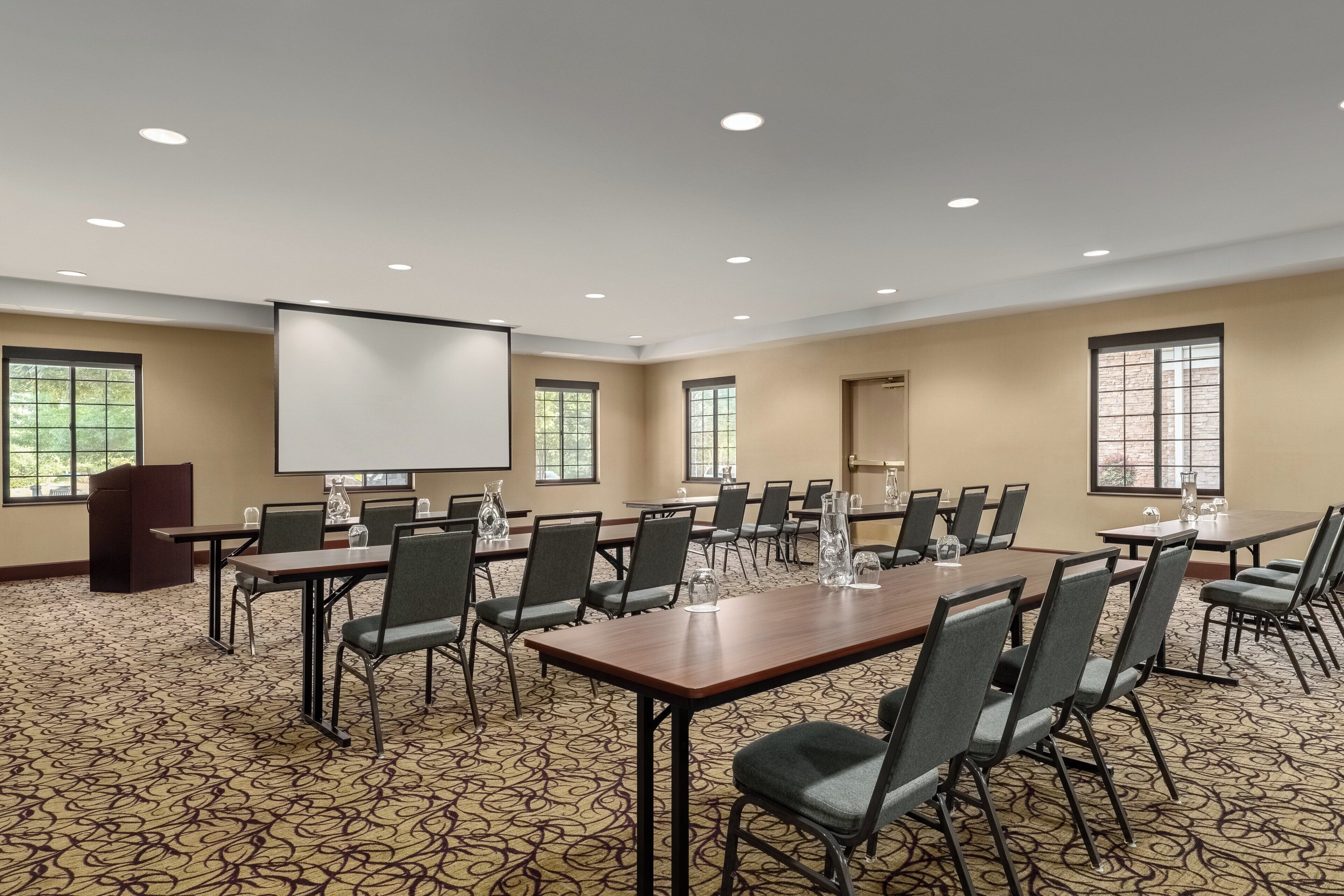 Un salón de conferencias con una gran mesa, varias sillas, una pantalla de proyector y ventanas.