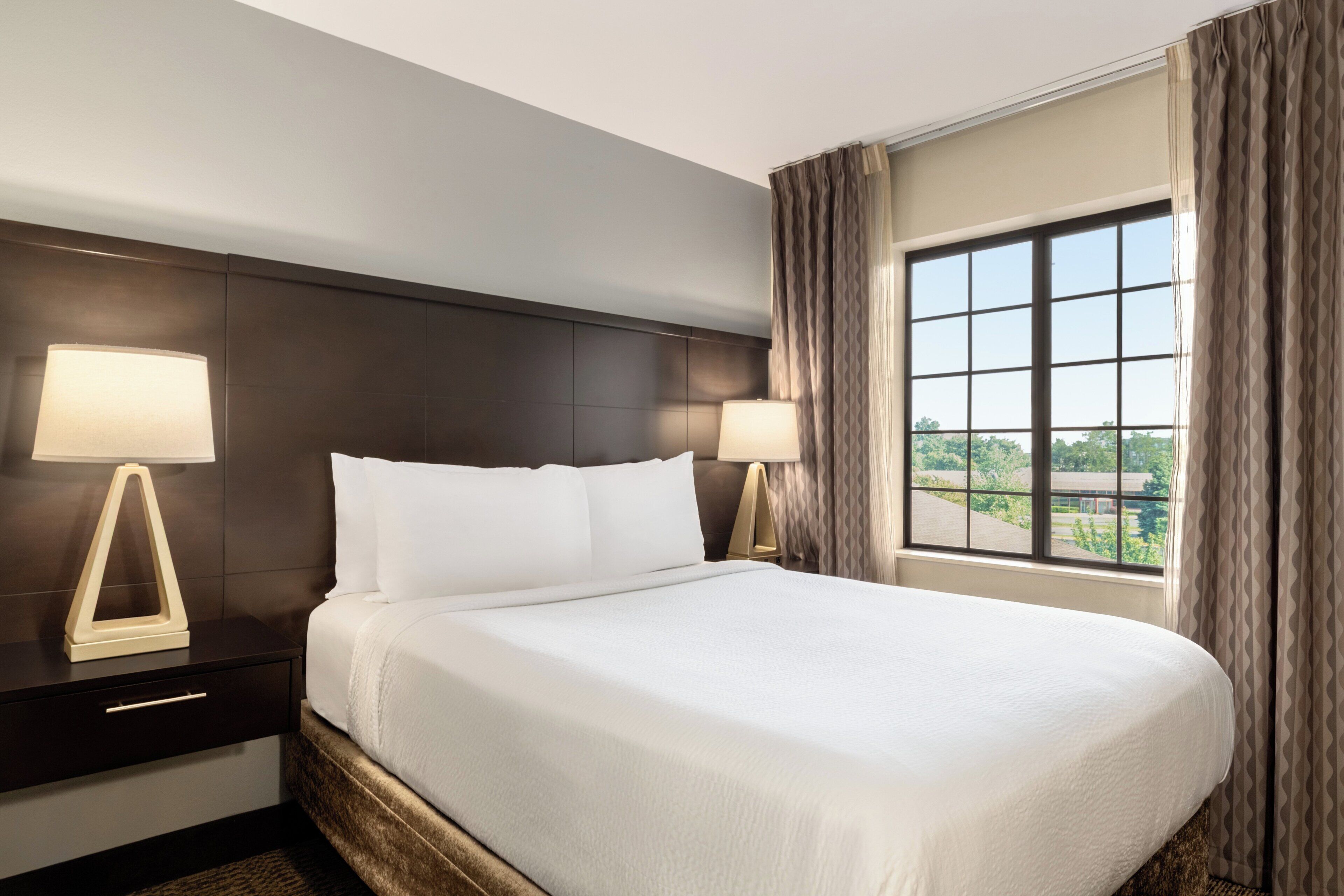 A hotel room with a large bed, two bedside tables, a window with curtains, and a view of greenery outside.