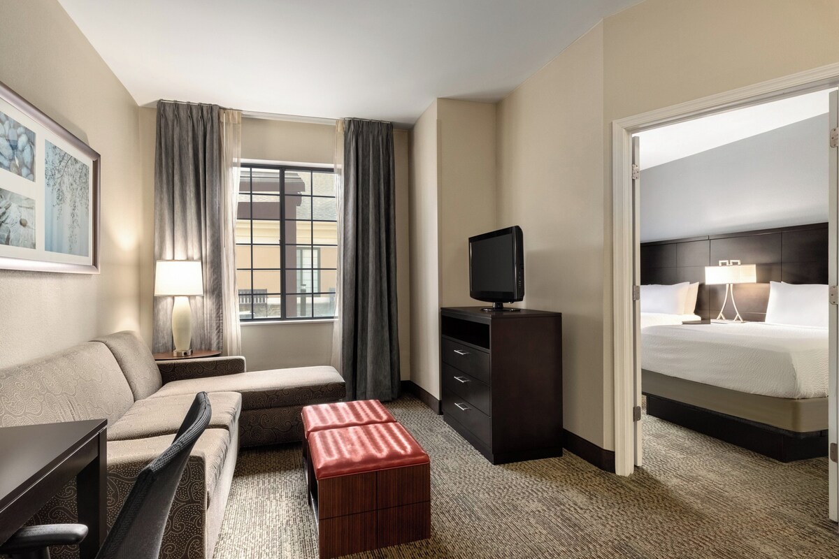 A hotel room with a sofa, a small ottoman, a television on a stand, a bed with white bedding, and a window with curtains.