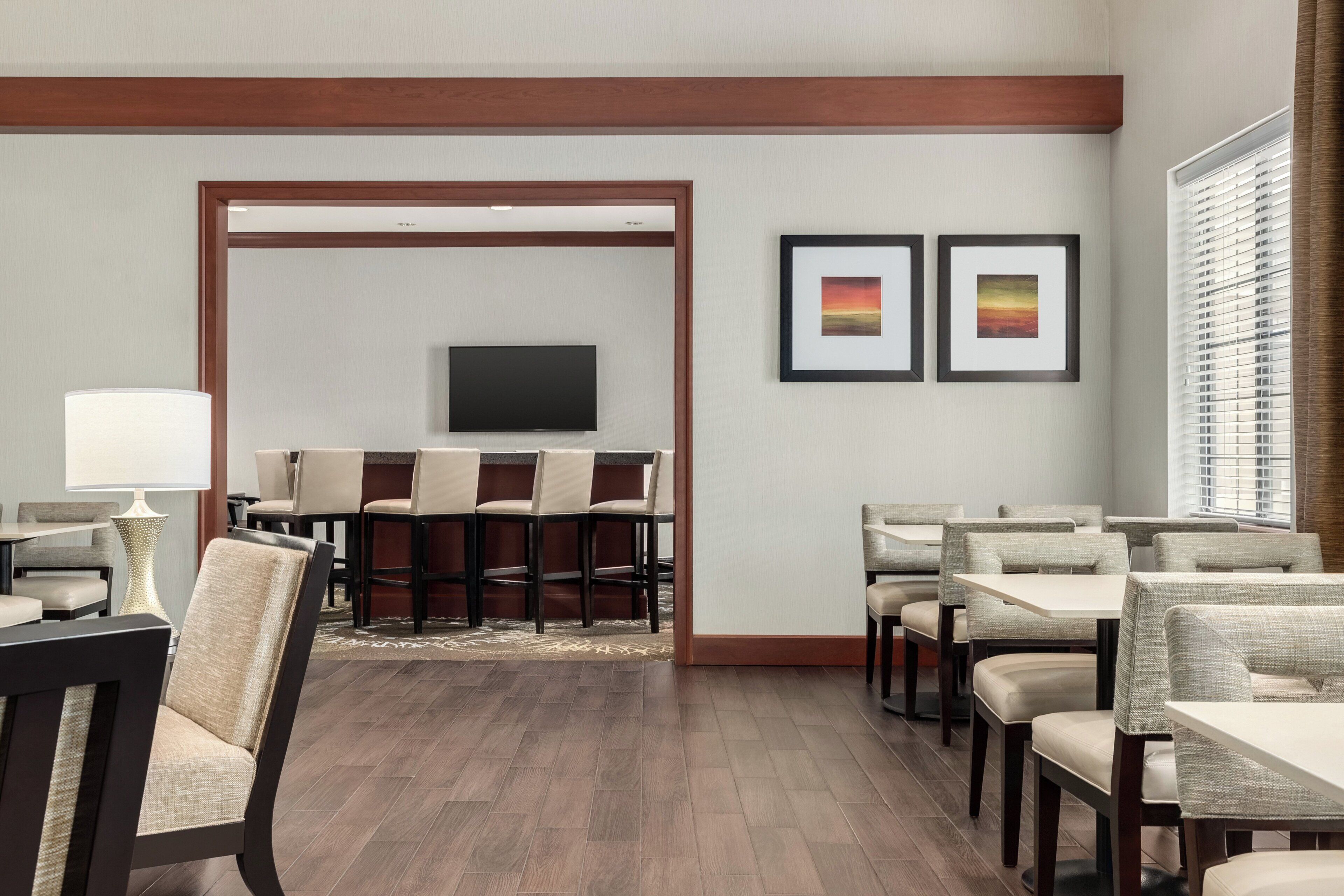 A hotel dining area with a TV mounted on the wall, framed artwork, and a view of the outside through the window.