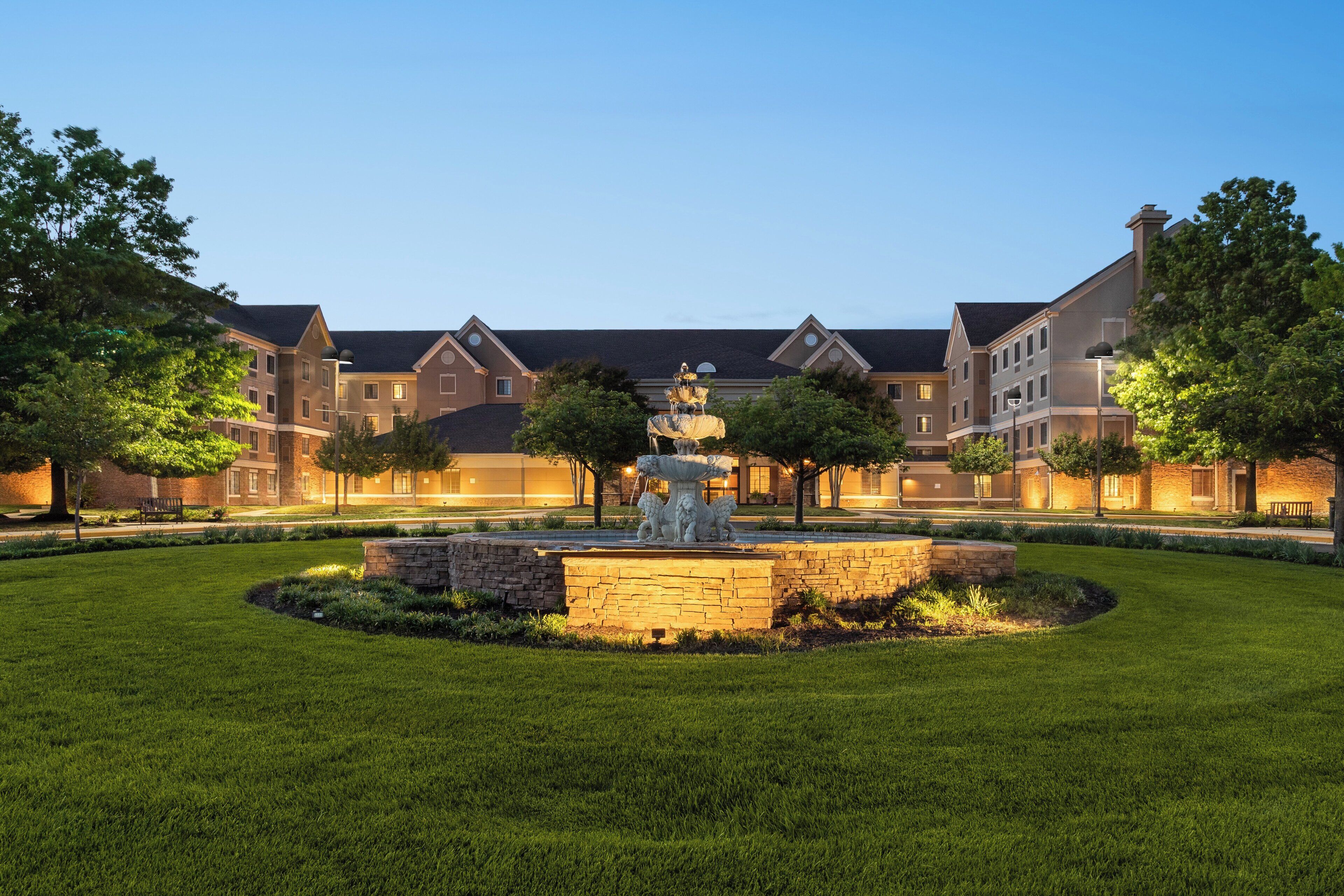A courtyard with a fountain, surrounded by well-maintained grass and trees, and a building with multiple windows and a brick facade.