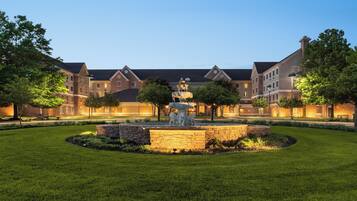 A courtyard with a fountain, surrounded by well-maintained grass and trees, and a building with multiple windows and a brick facade.
