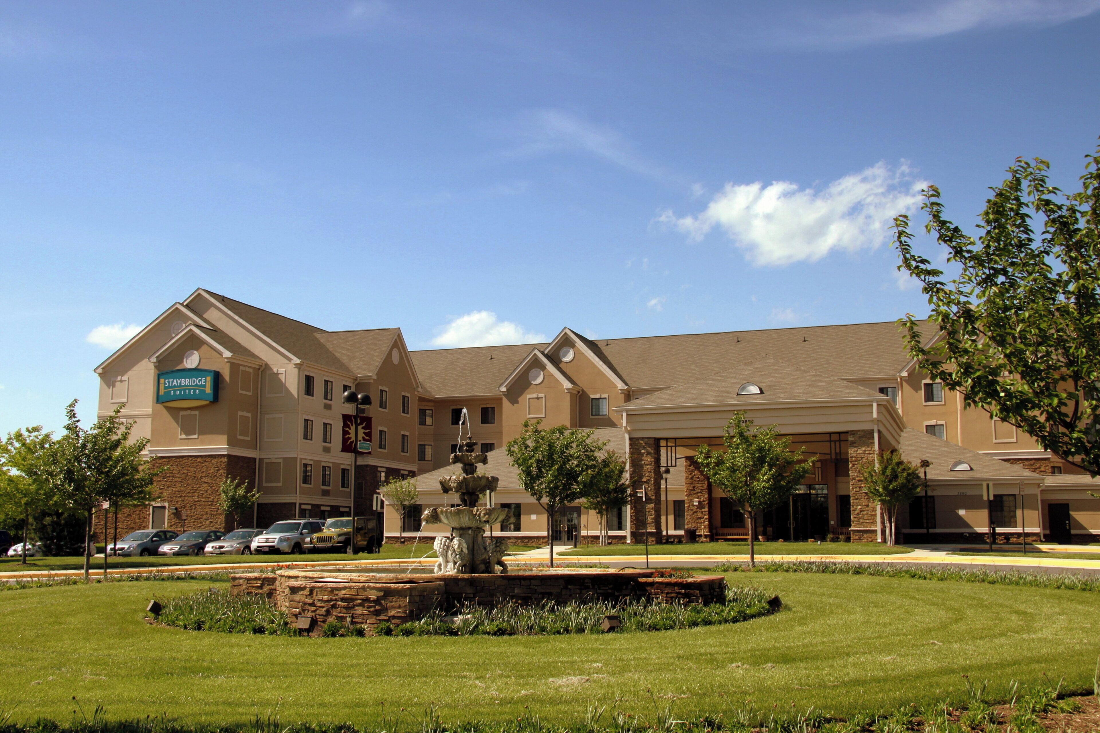 A Staybridge hotel with a central courtyard featuring a fountain, surrounded by well-maintained lawns and trees.
