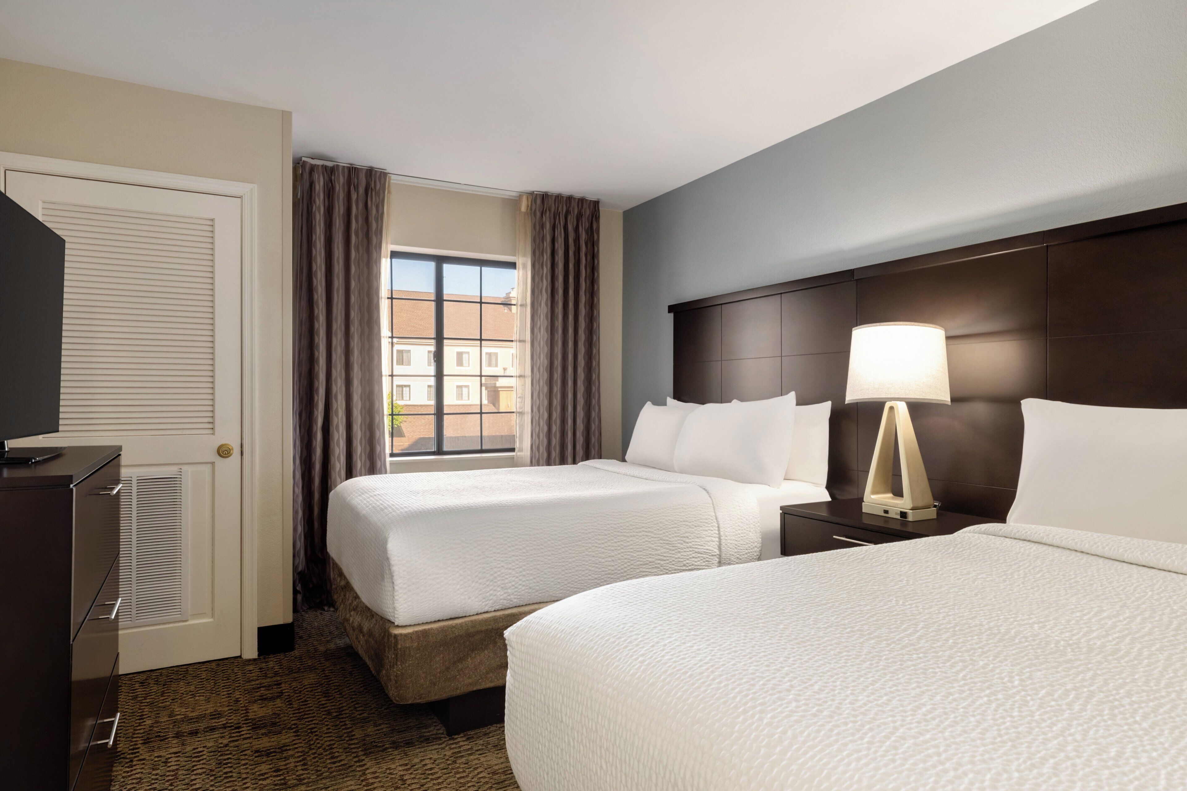 A hotel room with two beds, a desk, a lamp, and a view of buildings outside the window.