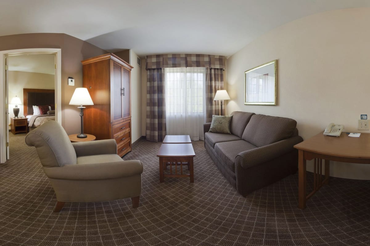 A hotel room with a sofa, armchair, small table, and a desk with a telephone. There is a wardrobe, a lamp, and a window with curtains.