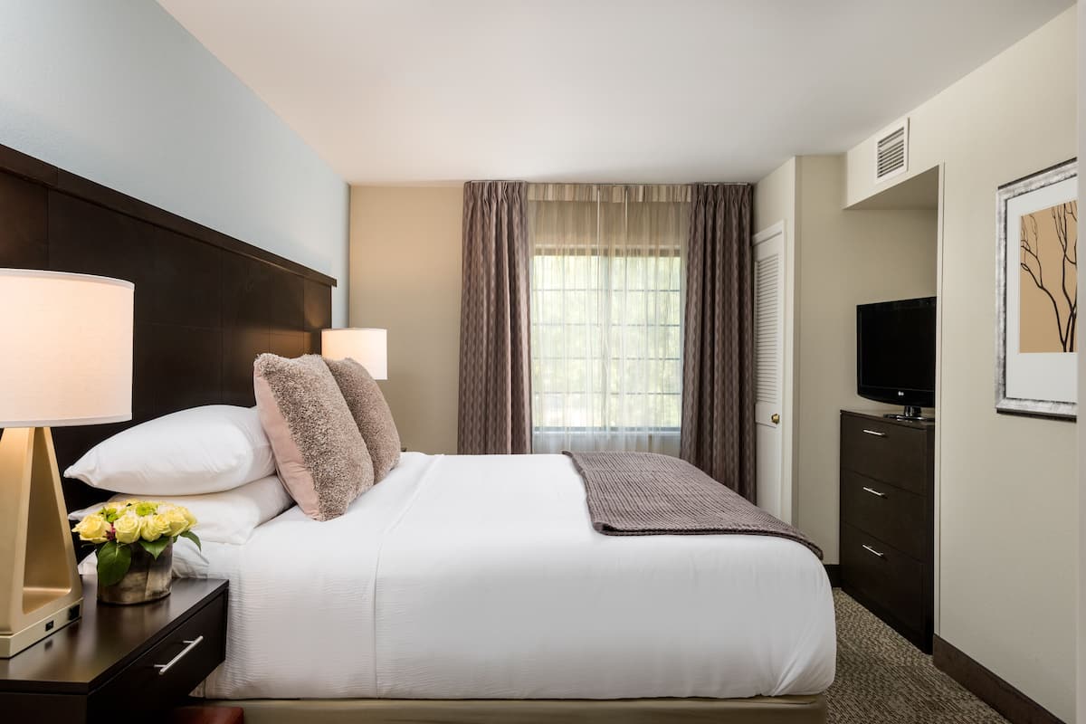 A hotel room with a large bed, two bedside lamps, a TV, a dresser, and a window with curtains.
