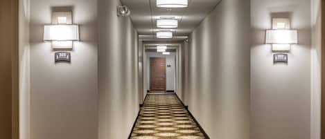 Un pasillo de hotel con piso alfombrado, luces empotradas en la pared y números de puerta.