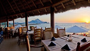 Ein Essbereich im Freien mit Tischen und Stühlen mit Blick auf einen Strand und einen Sonnenuntergang.