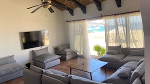Ein Wohnzimmer mit Fernseher, Deckenventilator und großen Fenstern mit durchsichtigen Vorhängen.