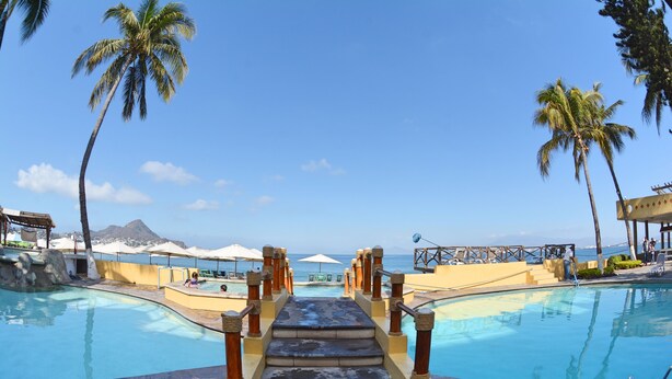 Ein Poolbereich mit einer Treppe zum Strand, umgeben von Palmen und Sonnenschirmen.